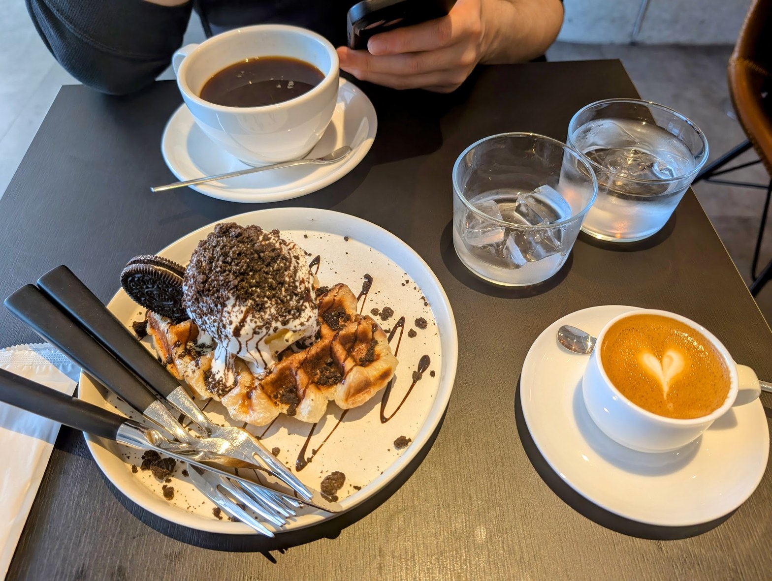 ワッフルとオレオクリームのデザートプレート、ラテアートのカフェラテ、コーヒーカップ、グラスが黒いテーブルに並ぶ