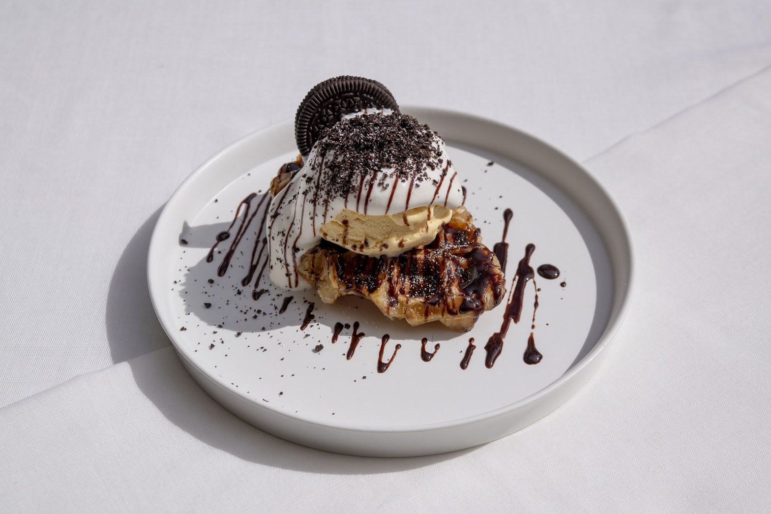 白い丸皿にのったワッフル、クリームとオレオクッキーとチョコソースを重ねたデザート、白い背景に自然光