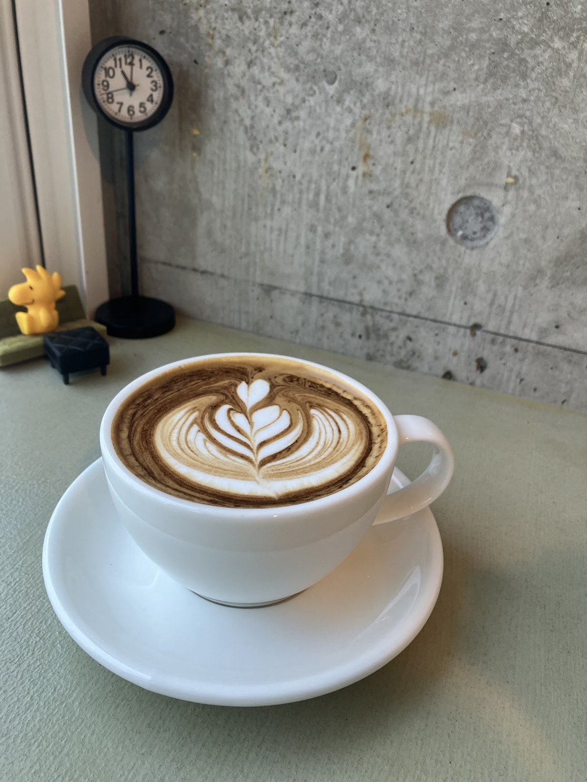 白いカップ＆ソーサーに注がれたラテアートのカフェラテ、奥にコンクリート壁と小型時計とスヌーピーのフィギュア